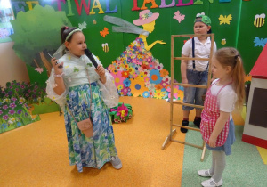Dziewczynka stoi z mikrofonem skierowana w kierunku dziewczynki. Z boku przygląda się chłopiec. W tle dekoracja lasu.